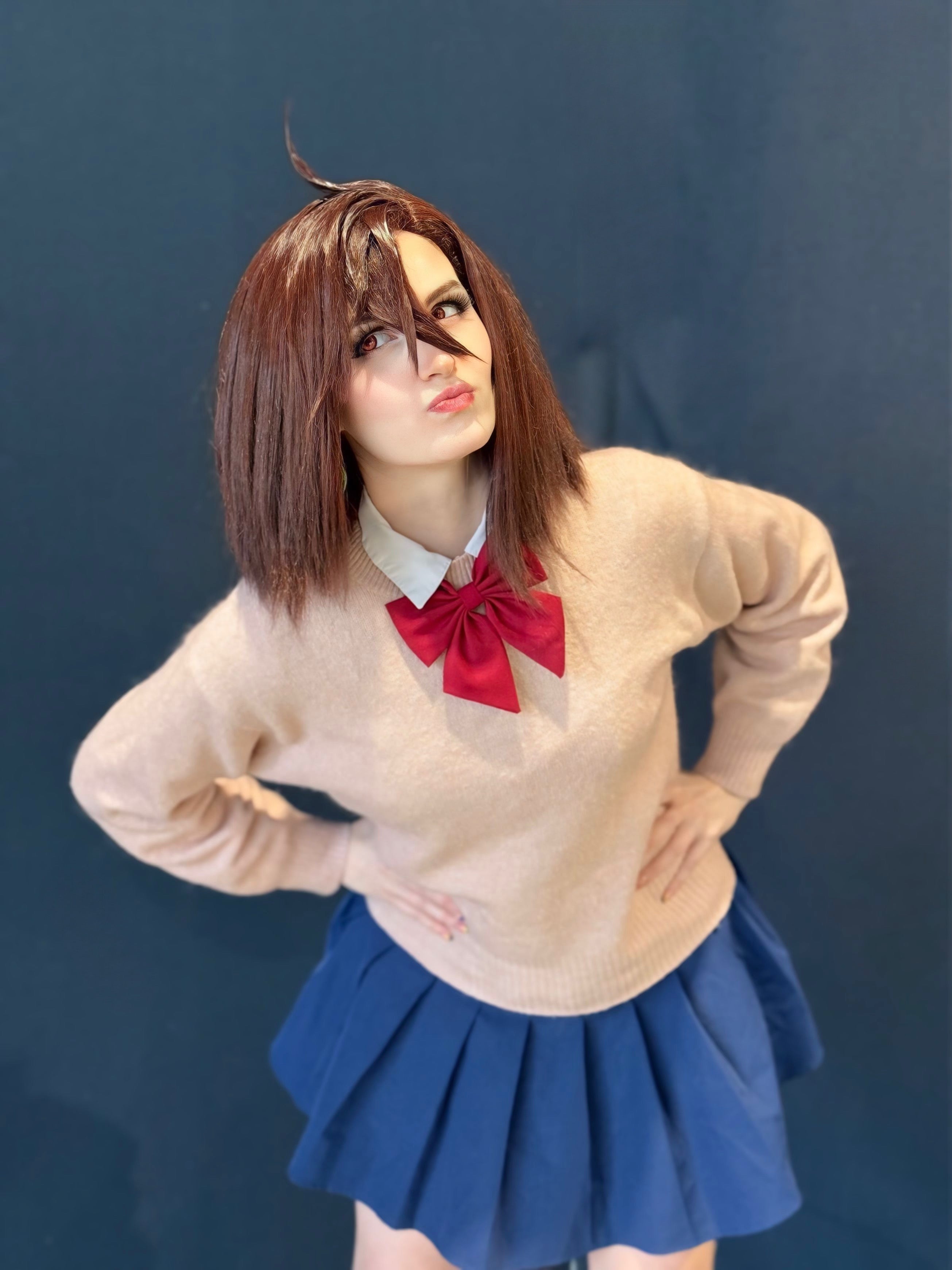 A cosplayer modeling the dark brown Momo Ayase cosplay wig from Dandadan, wearing her signature school uniform with a tan sweater, red bow, and blue pleated skirt to show the final styled look.